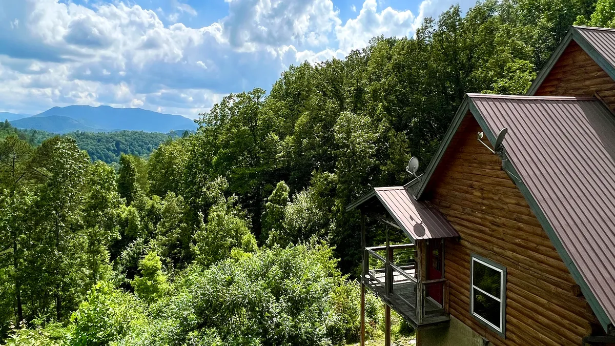 The image showcases a charming log cabin nestled among lush greenery, with majestic Smoky Mountain peaks rising in the background under a dramatic cloudy sky. The inviting front porch promises peaceful moments, and the natural setting hints at outdoor adventures and quiet relaxation.