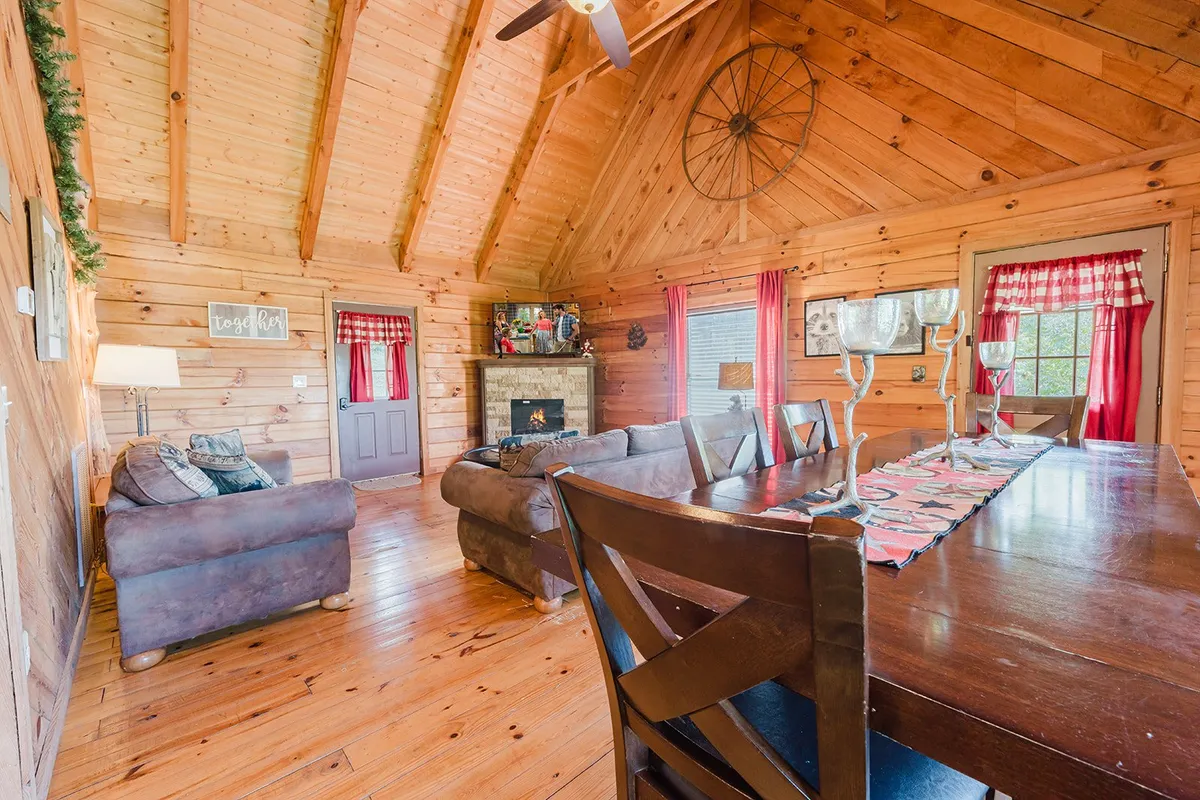 This image captures a spacious living and dining area with a rustic wooden ceiling, a cozy fireplace, a dining table set for group meals, and a comfortable sitting area. Large windows let in natural light and offer a glimpse of the outdoors, creating a welcoming space for groups to gather and socialize.