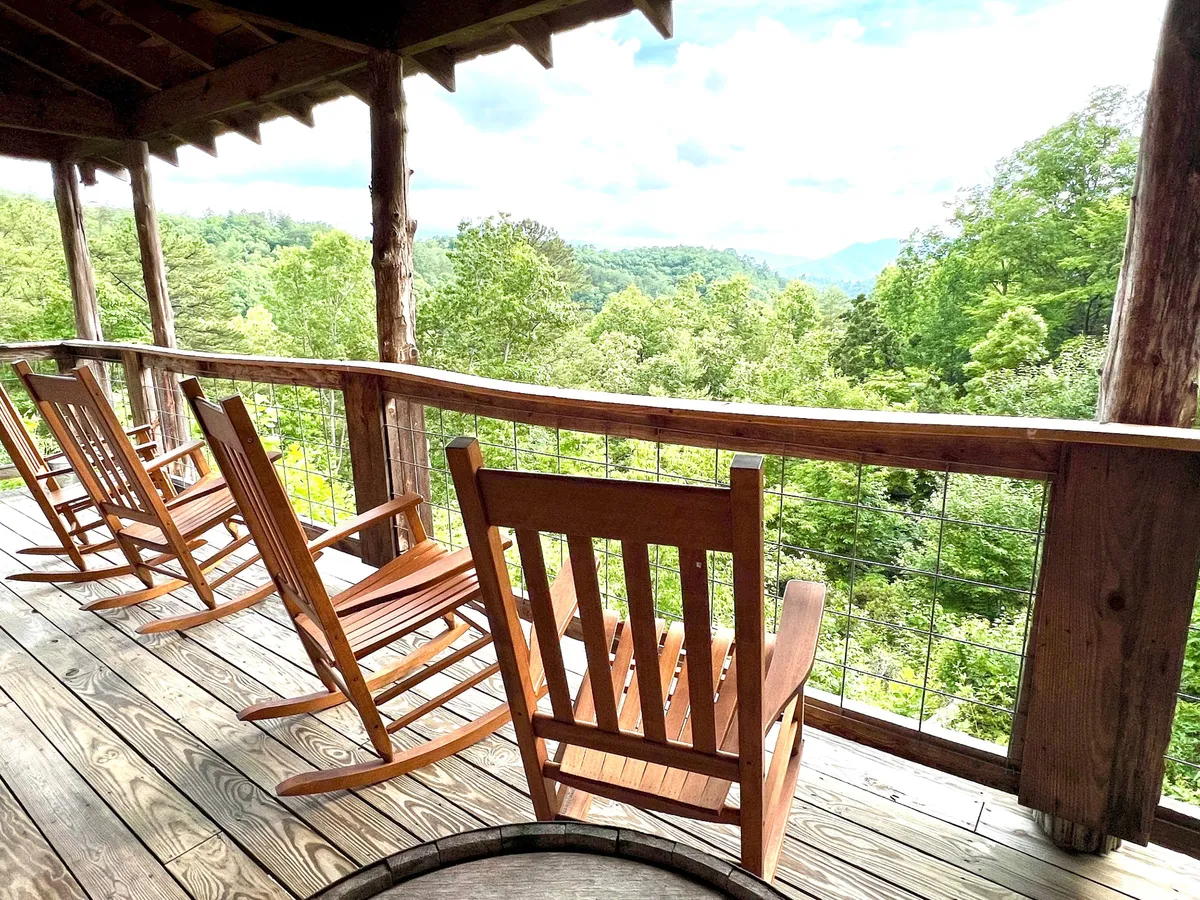 The image shows a pair of classic wooden rocking chairs positioned on a covered deck, framed by a sturdy wooden railing. Beyond the deck, a sweeping mountain view unfurls, lush greenery blending into distant summits. The scene is peaceful and intimate, perfect for enjoying quiet moments together.
