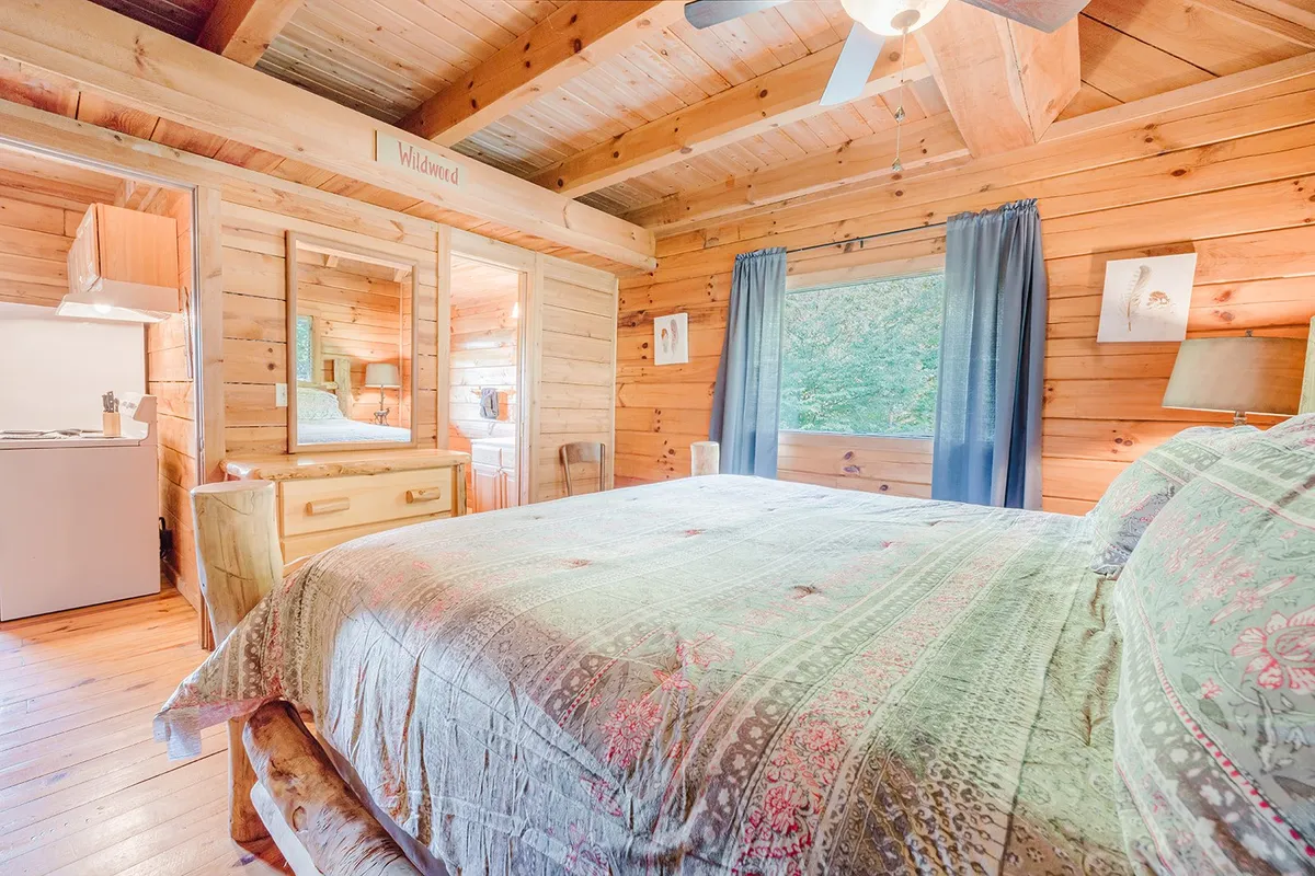 Inside, a beautifully made bed with a patterned quilt is bathed in natural light streaming through a large window. Wooden furniture and warm accents create a tranquil, inviting space perfect for unwinding after a day of exploration.