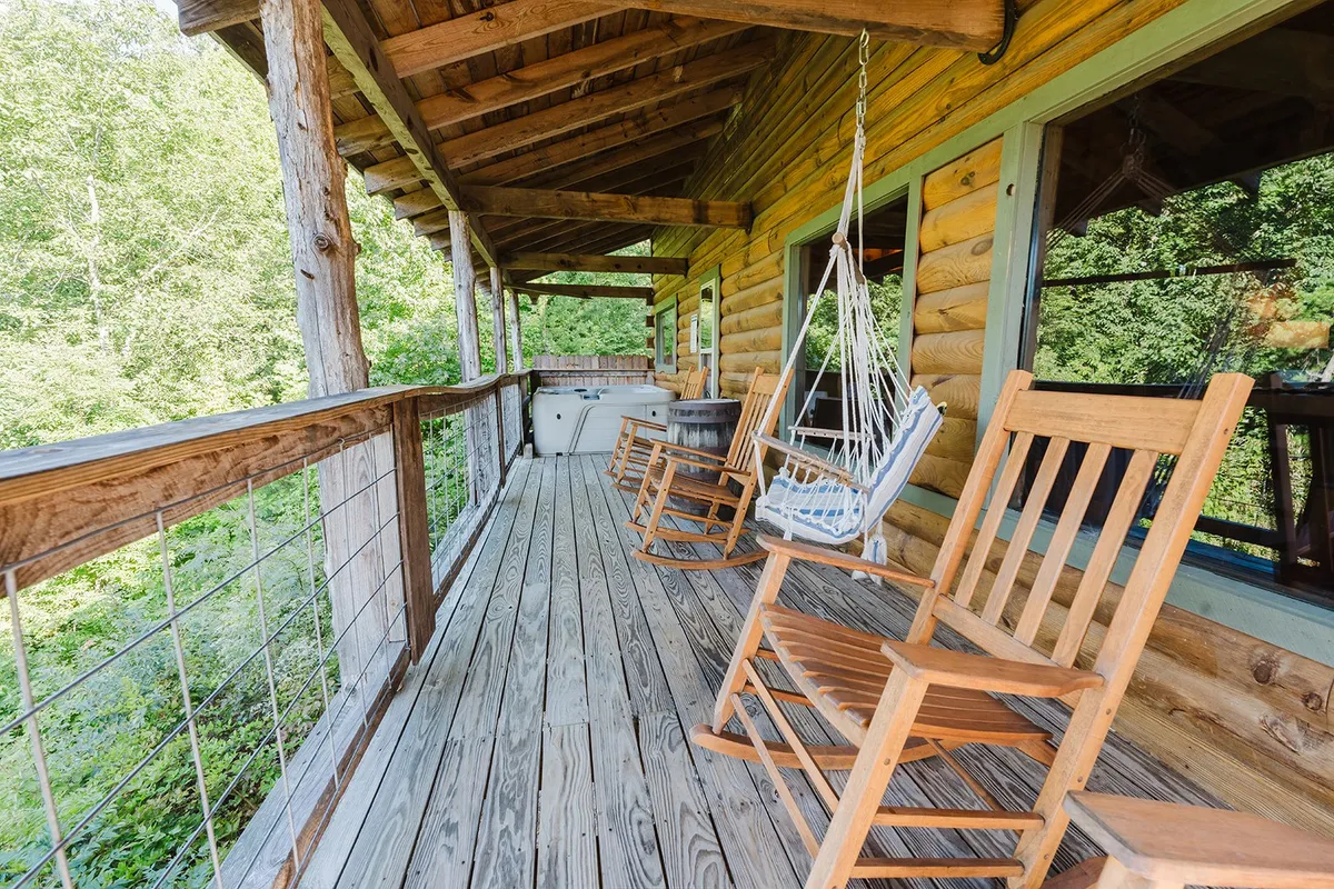 This exterior shot provides a wide view of the covered deck, highlighting wooden rocking chairs, a hammock, wooden railing, and the surrounding greenery. The serene setting invites moments of quiet reflection, reading, or enjoying time with loved ones.