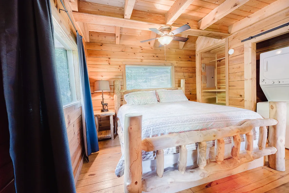 This image features a log bed against warm wooden walls, a window with curtains bringing in soft natural light, and a ceiling fan overhead. The inviting bedding and intimate setting evoke a sense of peaceful seclusion—ideal for restful nights and private retreats.