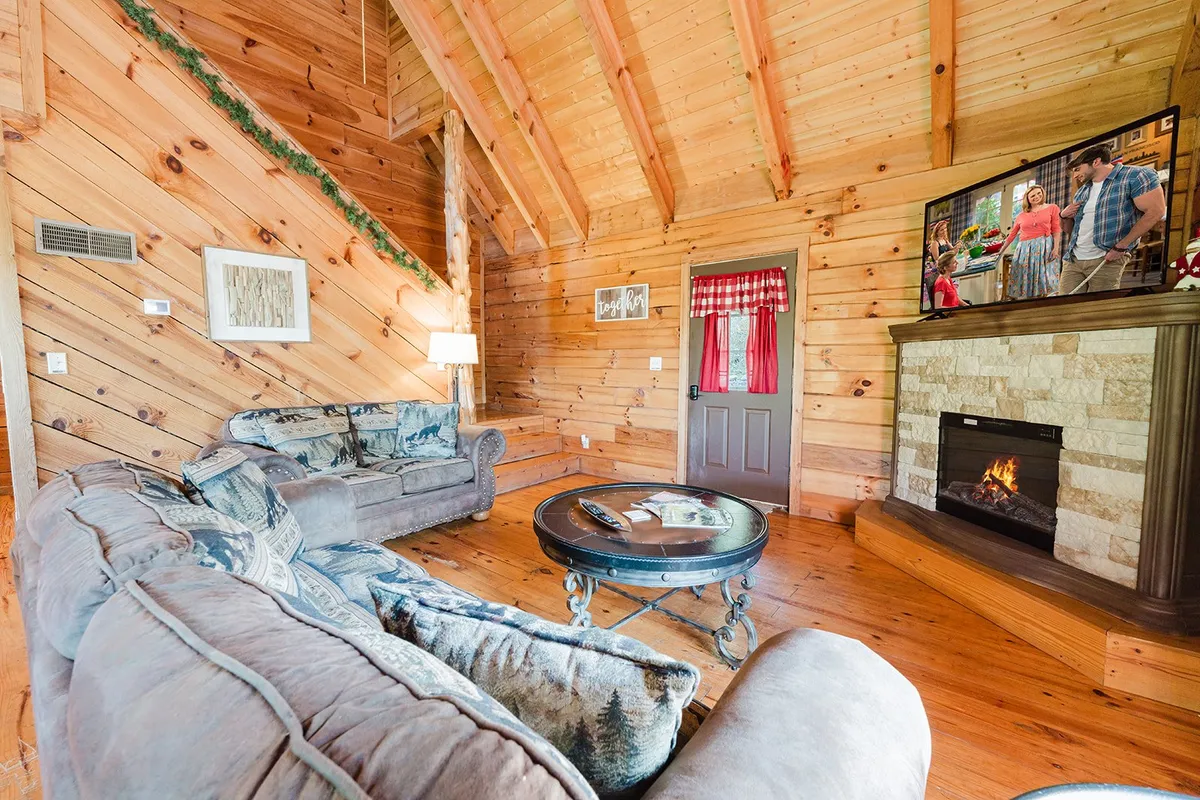 A warmly lit living room featuring plush sofas gathered around a glowing electric fireplace, with wooden walls lending rustic charm. Large windows allow natural light to stream in, illuminating the inviting space. The setup encourages relaxation, conversation, and gatherings with friends, family, or pets.