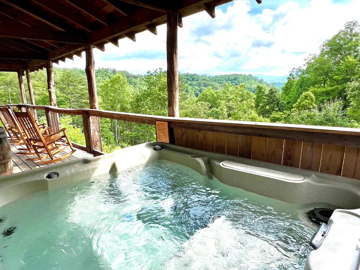 This exterior image showcases a covered deck with a bubbling hot tub, wooden rocking chairs, and a breathtaking view of the mountains. The scene is tranquil, inviting relaxation and social connection amidst stunning natural surroundings.