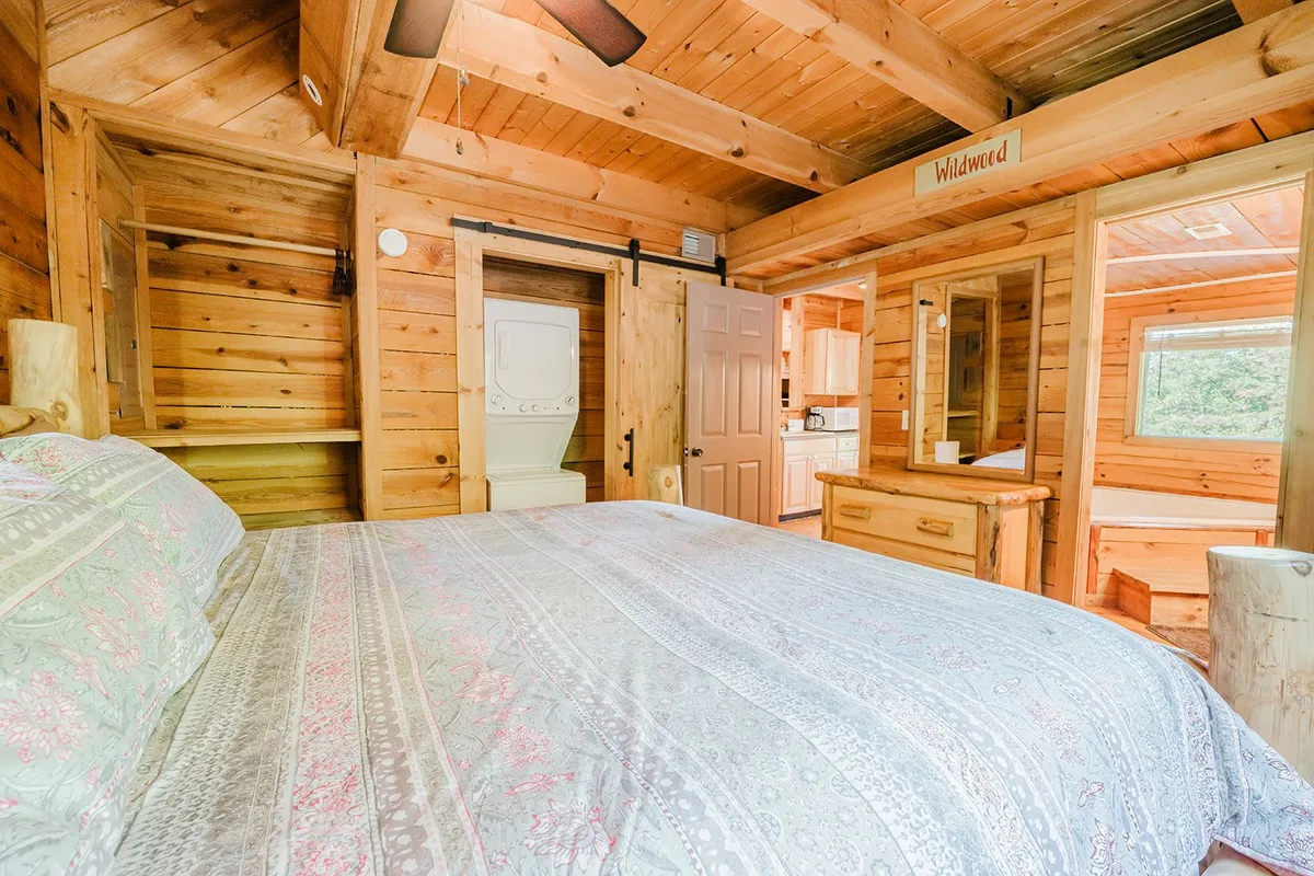 The scene highlights a bed with patterned bedding, natural wood paneling, and a window that fills the room with daylight. Wooden furniture and tranquil decor create a comfortable and restful environment, ideal for a guest or upstairs bedroom.