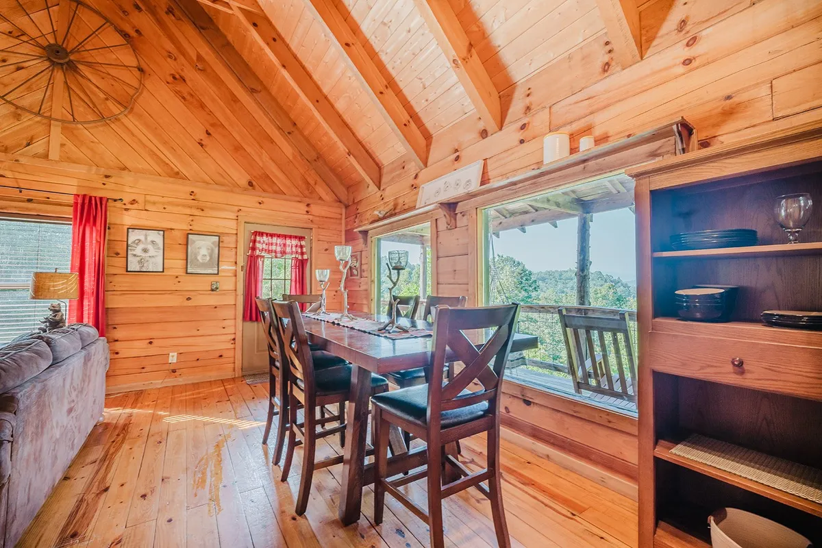 The photo features a spacious dining table for six, framed by large windows that let in scenic natural light. The warm wood ceiling and shelves with dishware create a cozy yet open atmosphere. The table is set for family meals, game nights, or planning sessions.